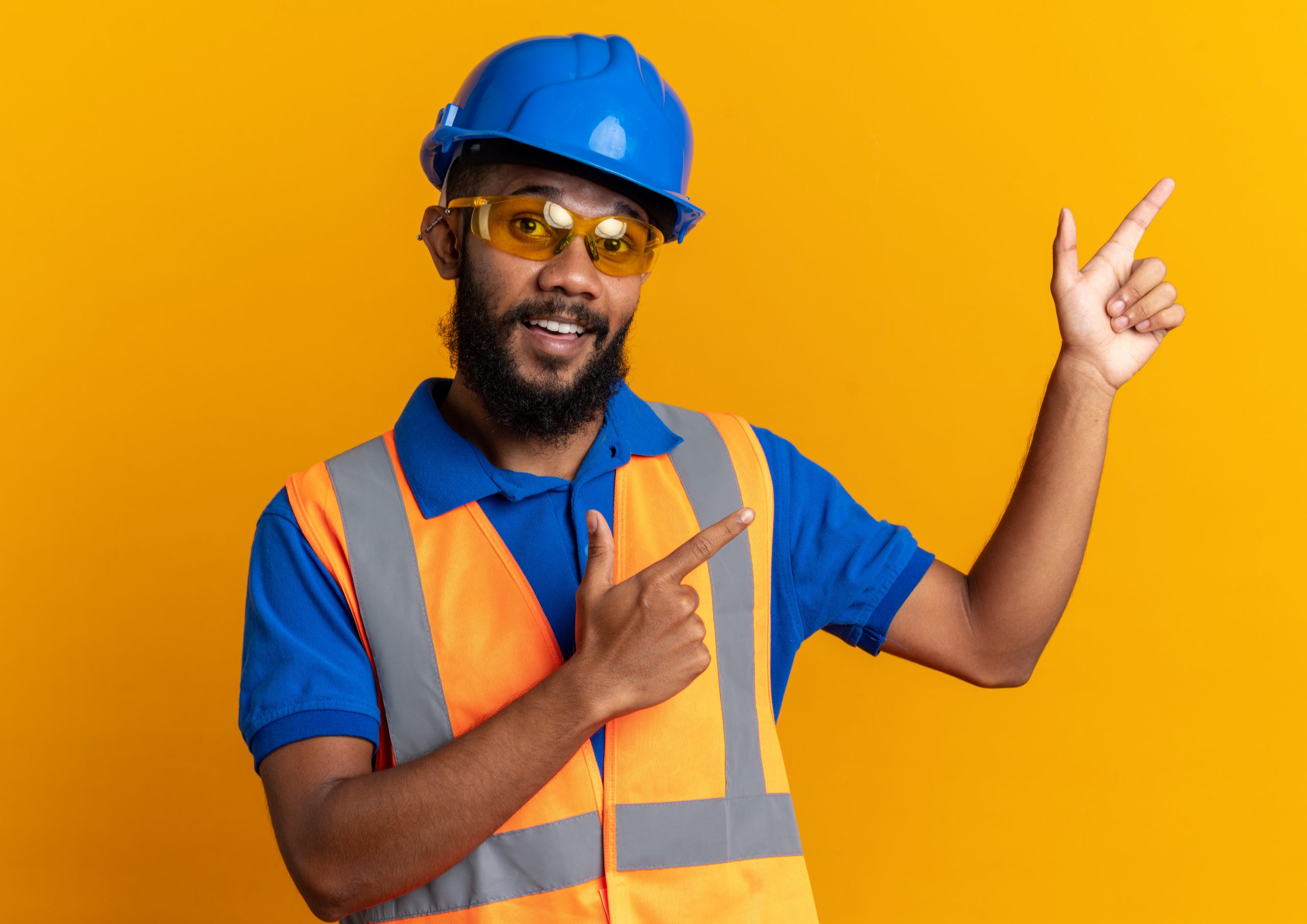 impressed young afro-american builder man in safety glasses wearing uniform with safety helmet pointing at side isolated on orange background with copy space