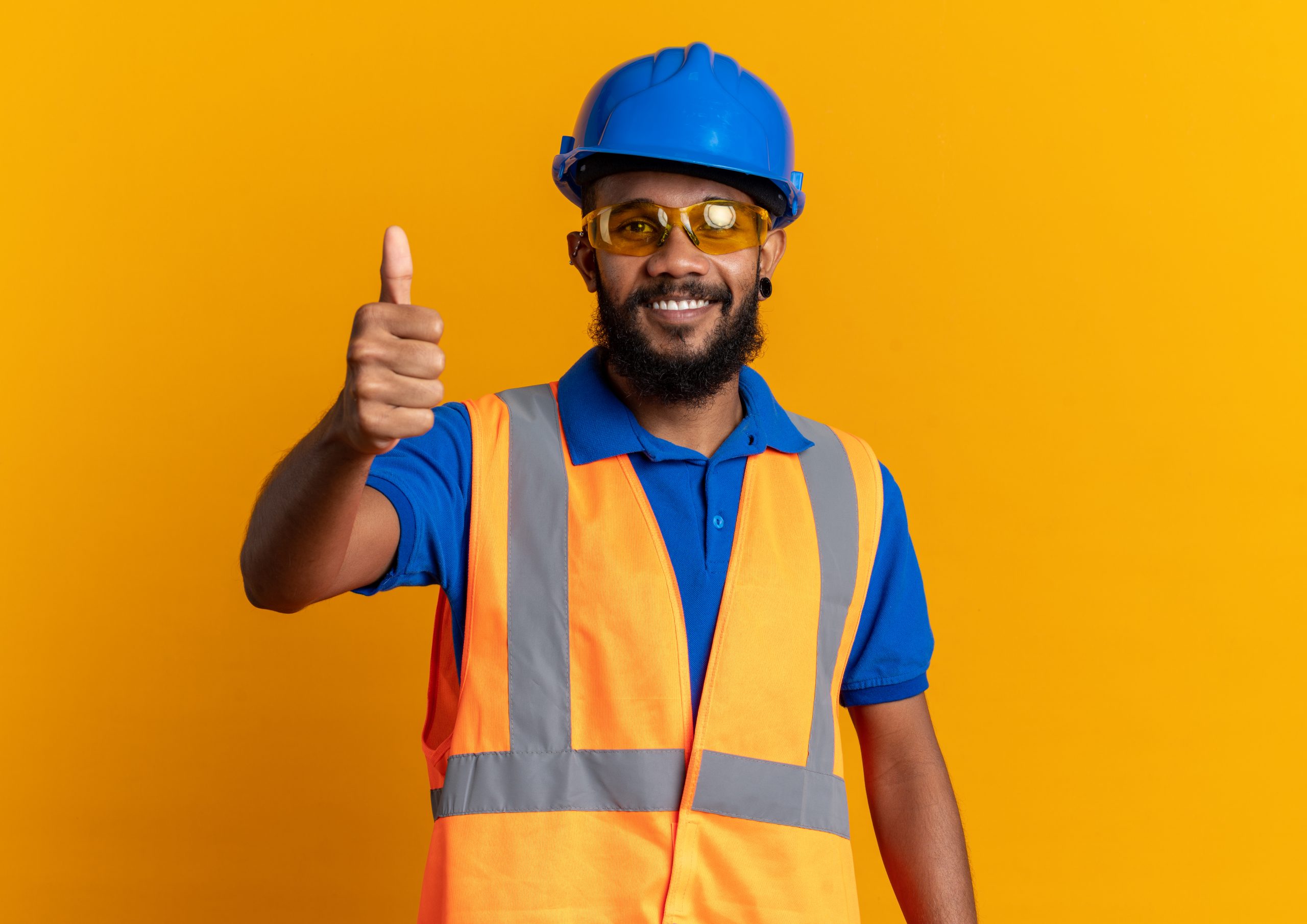 smiling young afro-american builder man in safety glasses wearing uniform with safety helmet thumbing up isolated on orange background with copy space