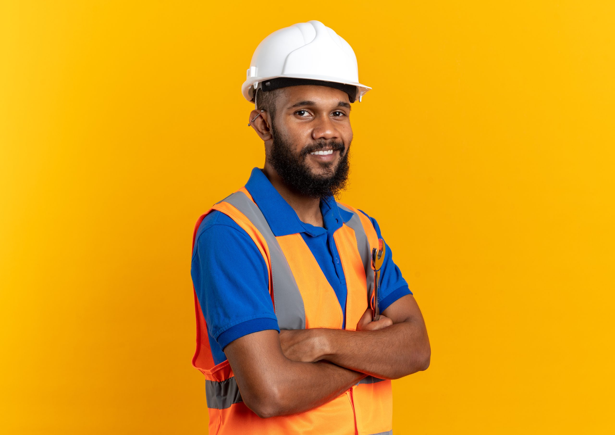 smiling young afro-american builder man in uniform with safety helmet standing with crossed arms isolated on orange background with copy space